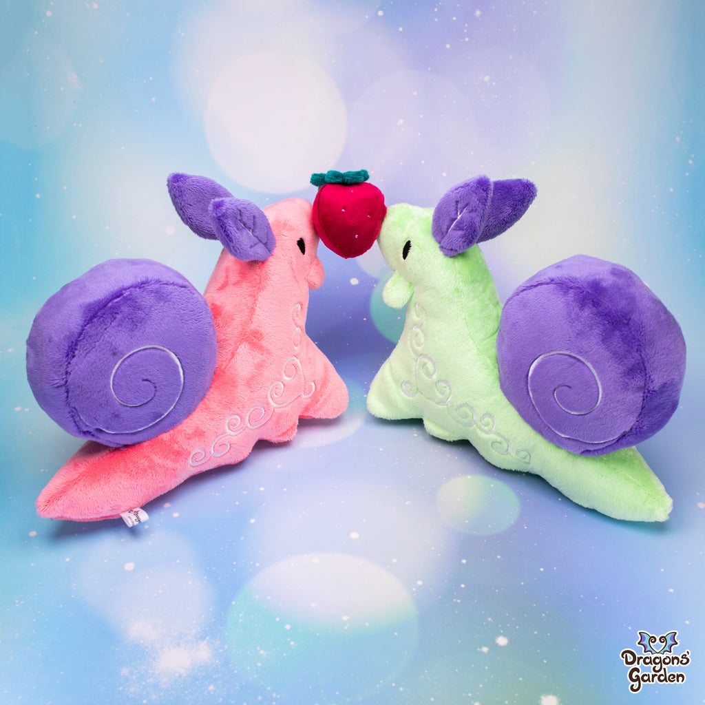 pink and green snail plushies with purple shells holding up a plush red strawberry