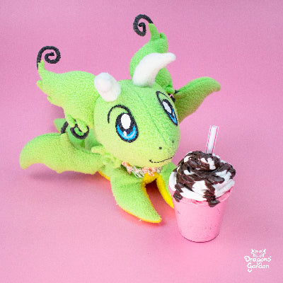 Manu the green dragon plushie ona pink background with a strawberry milkshake with cream and covered in chocolate