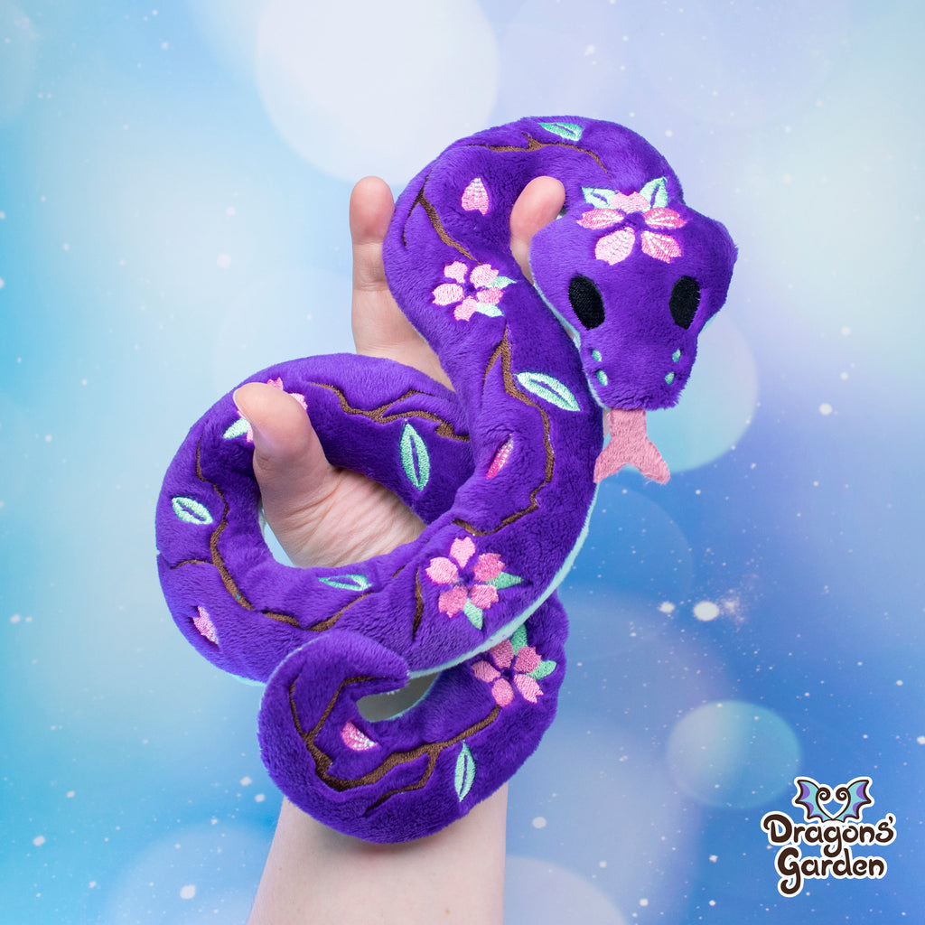 purple body with pink sakura cherry blossom snake plushie on a sparkly blue background being held with one hand